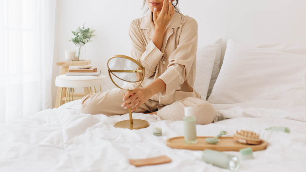 Mulher sentada sobre a cama, realizando a aplicação de creme facial. Há outros cosméticos sobre a cama. Ela está realizando um autocuidado.